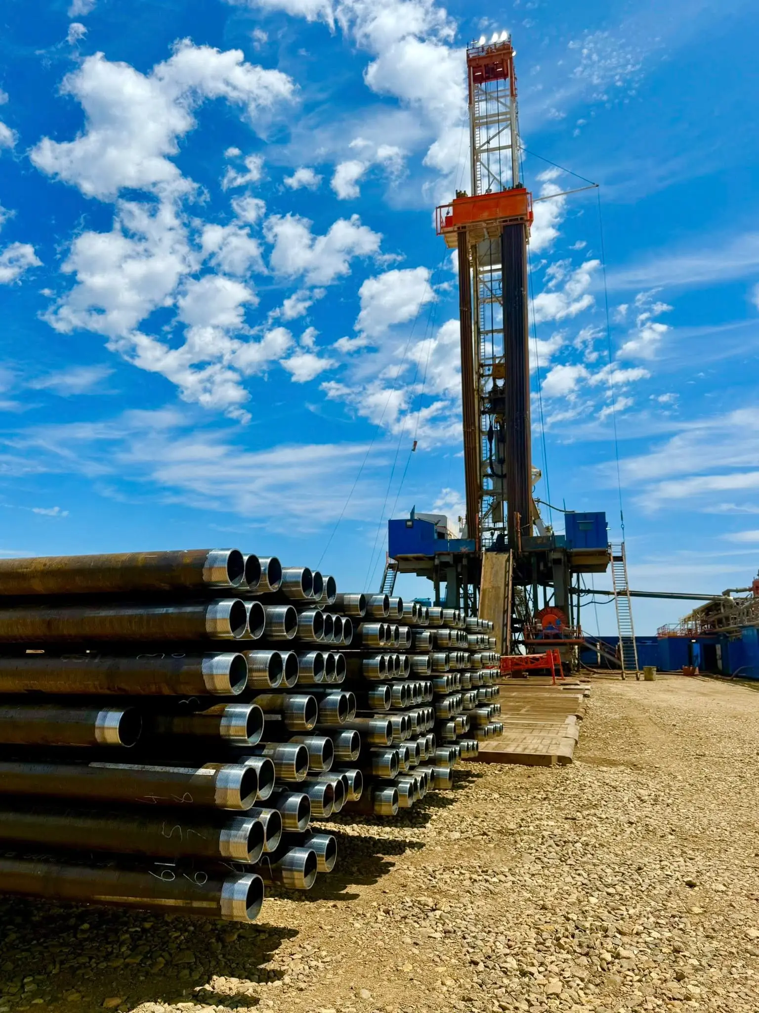 Stacked casing tubing in the foreground with a tall workover rig and clouds beyond