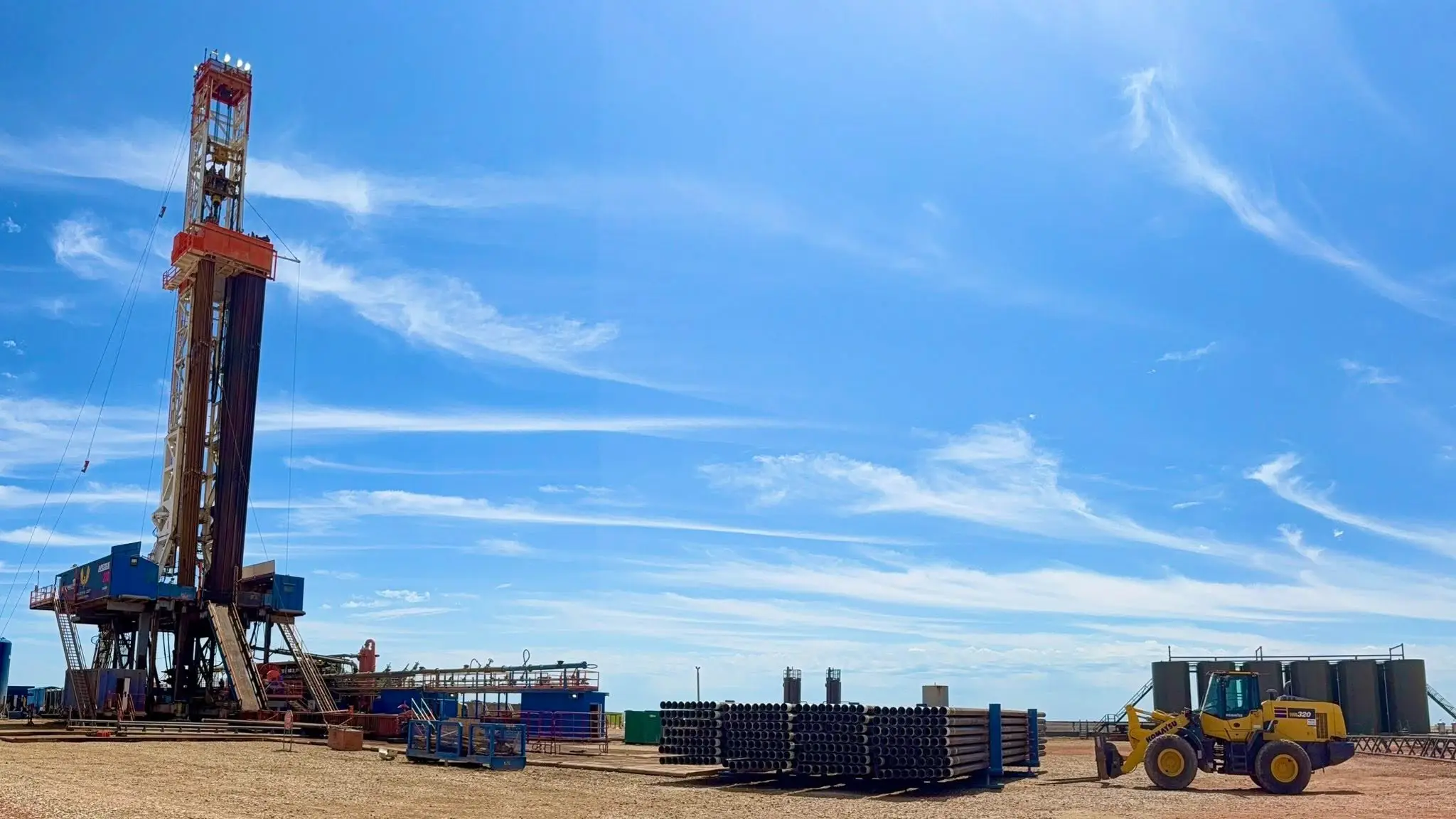 Wide view of a workover rig, Komatsu wheel loader, and stacked casing on a Bakken pad