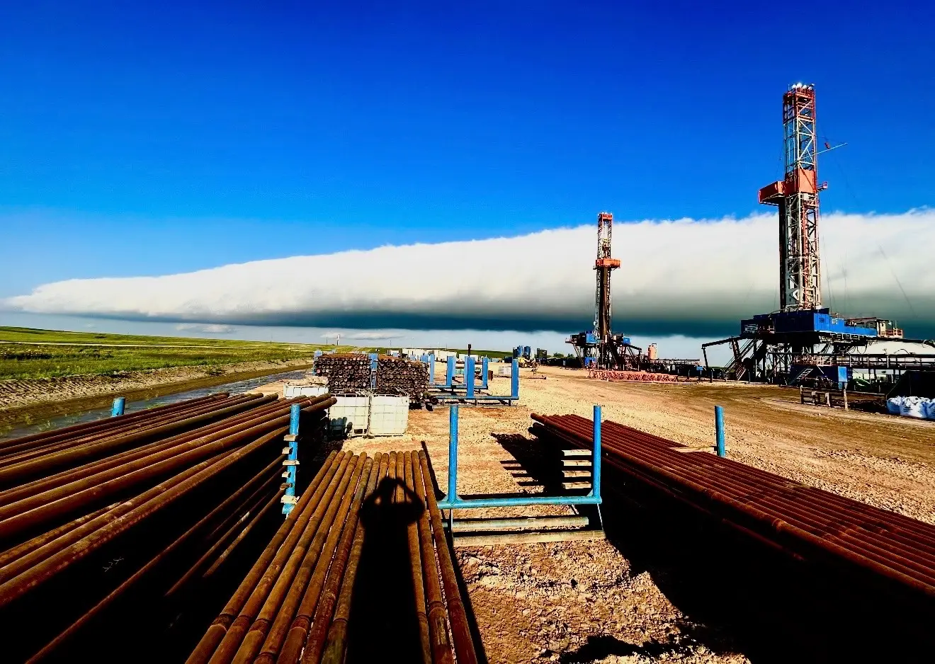 Two drilling rigs on the horizon with stacked drill pipe and a passing storm cloud over the Bakken